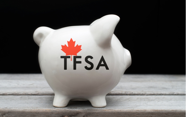 The side of a white piggy bank with the letters TFSA and a Canadian maple leaf printed on the side sitting on a wooden surface in front of a black background
