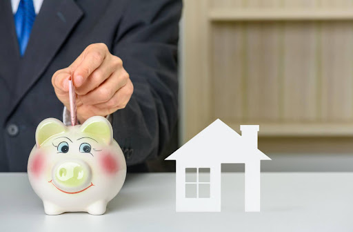 A close-up of a man putting $50 into a piggy bank and a model house next to it.