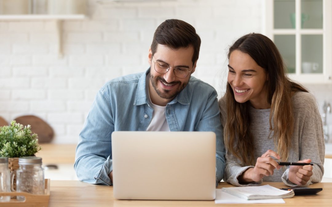 A young couple happily look over their laptop in a brightly lit kitchen organizing their finances.