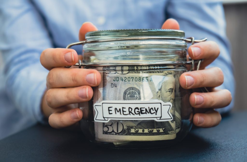 A close-up image of two hands holding a clear jar full of money with the label "EMERGENCY" scrawled on the front.