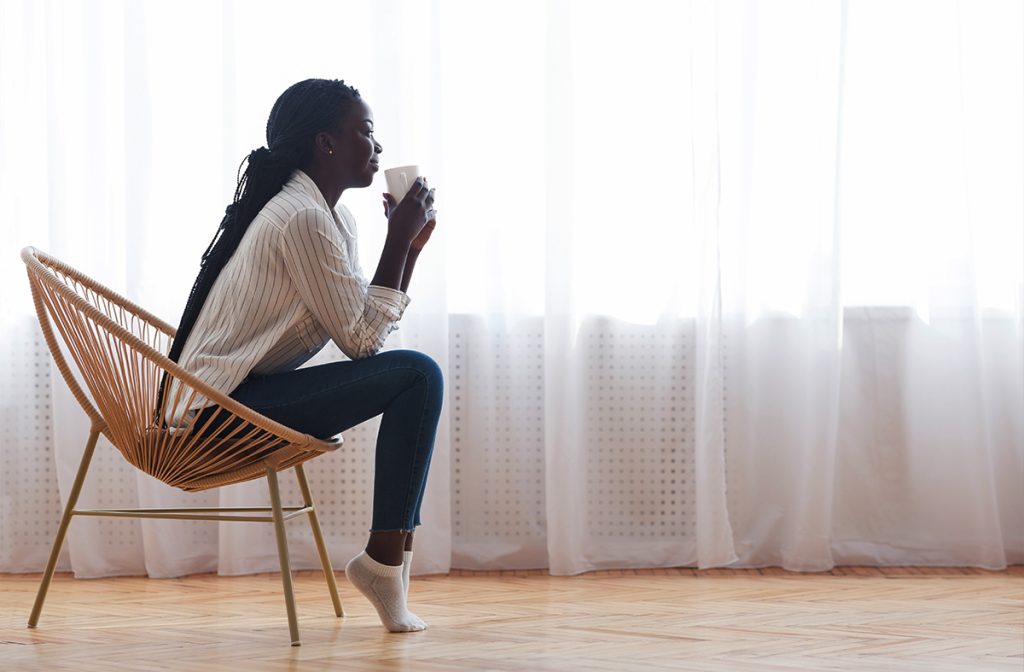 woman drinking a coffee in a front a a big bright window with curtains on it