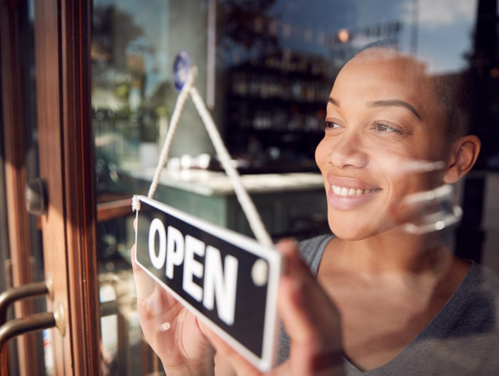 Female Owner Of Start Up Coffee Shop Or Restaurant Turning Round Open Sign On Door